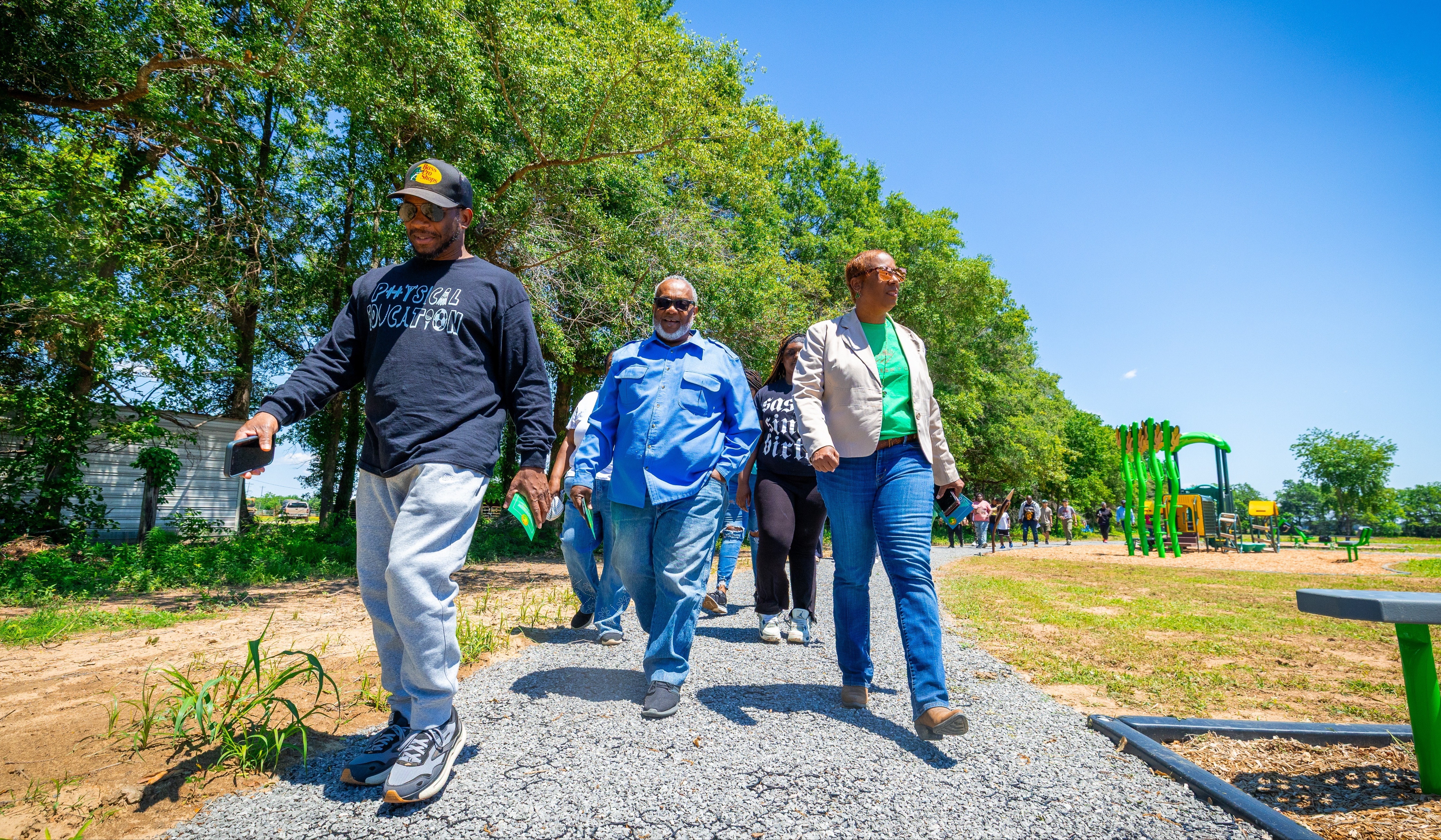 Group of adults walking on trail at park with playground equipment in background.l